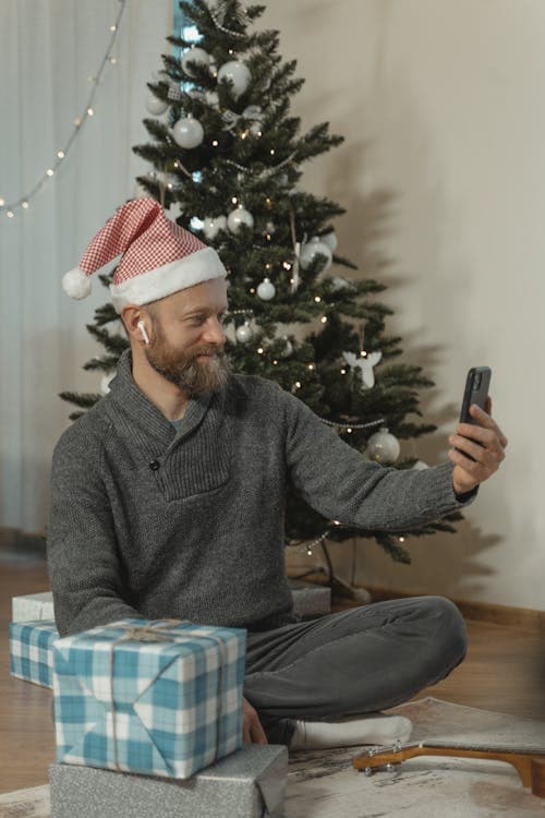 Man using smartphone with wireless earbuds near Christmas tree with gifts